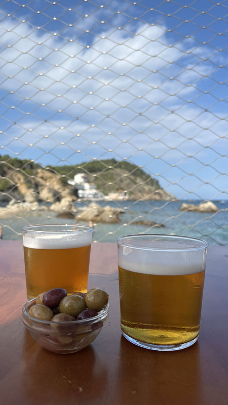 Ein Schälchen mit grünen Oliven und zwei Gläser gefüllt mir Bier auf einem Tisch, im Hintergrund ist das Meer zu sehen.