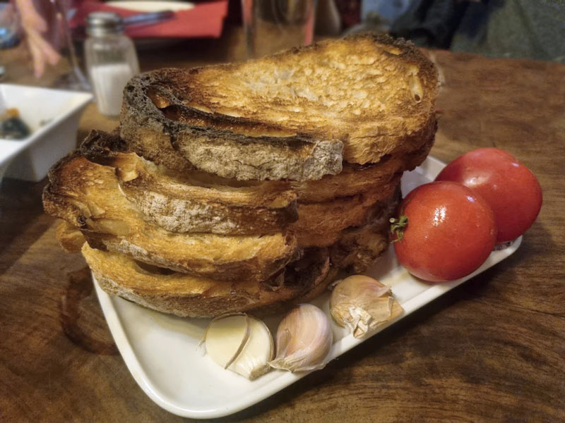 Aufeinandergestapelte und getoastete Brotscheiben auf einem Teller, serviert mit Tomaten und Knoblauchzehen.