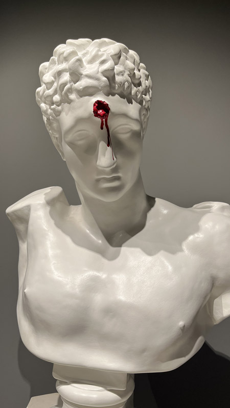 Römische, weiße Marmorstatue mit roter, blutender Wunde verursacht durch einen Kopfschuss.