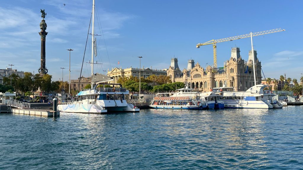 Blick vom Meer auf das Ufer: hinter ankernden Booten ragen die Kolumbussäule und das Portal de la Pau auf.